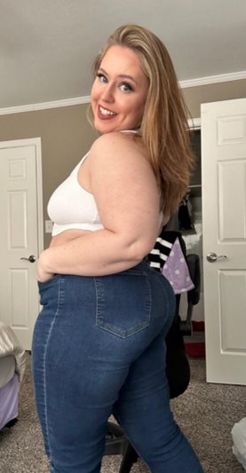Woman with blonde hair in white top and jeans posing in bedroom.