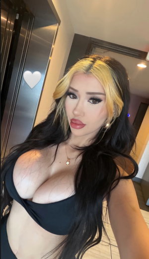 Woman with long black and blonde hair wearing black top, taking selfie indoors.
