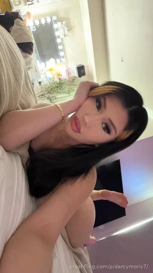 Woman with long dark hair and blonde highlights posing in a bedroom, wearing a black top and light