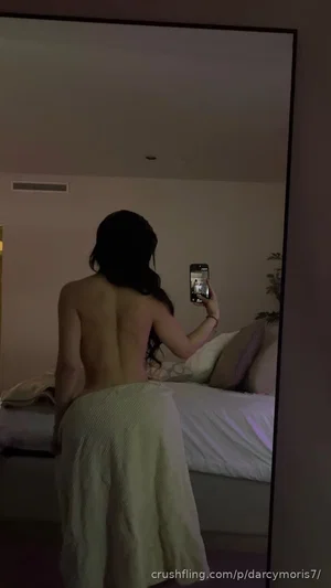 Woman with long dark hair taking mirror selfie, bare back, wrapped in white towel, in bedroom.