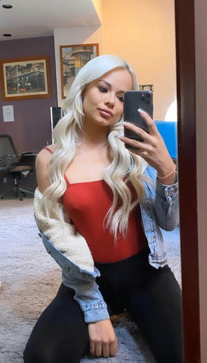 Woman with long platinum hair in red top and denim jacket, kneeling in room with carpet and office