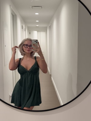 Woman with blonde hair and glasses in a green dress takes mirror selfie in a hallway.