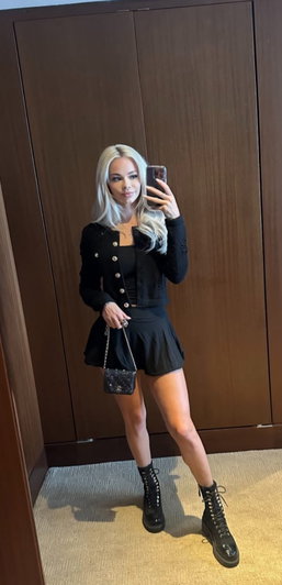 Blonde woman in black outfit and boots poses for mirror selfie in room.