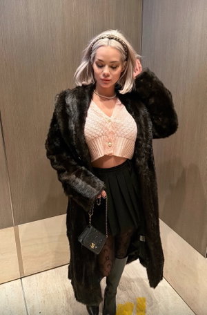 Woman with silver hair in black fur coat, pink crop top, black skirt, and patterned tights indoors.