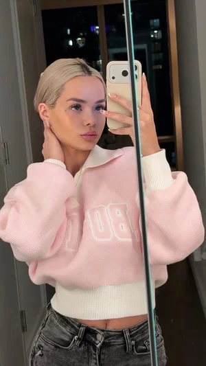 Woman with blonde hair in pink cropped sweater and dark jeans, taking mirror selfie indoors.