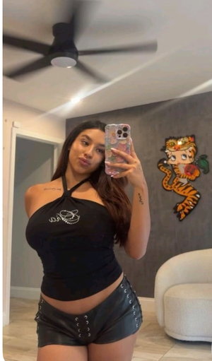Woman in black halter top and leather shorts takes mirror selfie in bedroom.