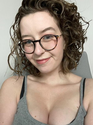 Woman with curly hair and glasses wearing a gray tank top, smiling at camera indoors.
