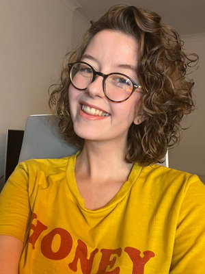 Woman with curly hair and glasses wearing a yellow t-shirt, smiling in a casual indoor setting.