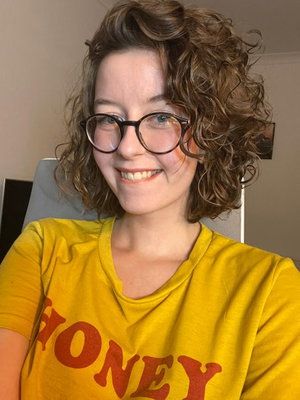 Woman with curly hair and glasses wearing a yellow t-shirt, smiling in a casual indoor setting.