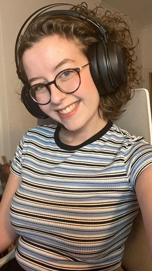 Woman with curly hair and glasses wearing striped shirt and headphones, smiling indoors.