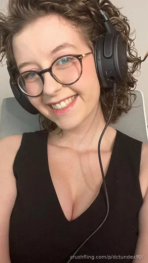 Woman with curly hair and glasses wearing black top and headphones, smiling indoors.
