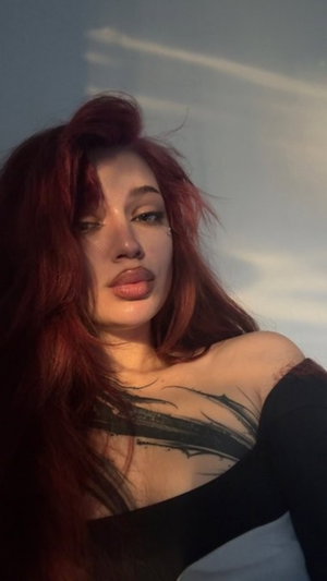 Woman with red hair and chest tattoo wearing black off-shoulder top, indoor setting.