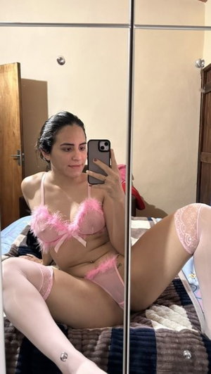 Woman in pink lingerie and stockings sitting on bed, taking mirror selfie.