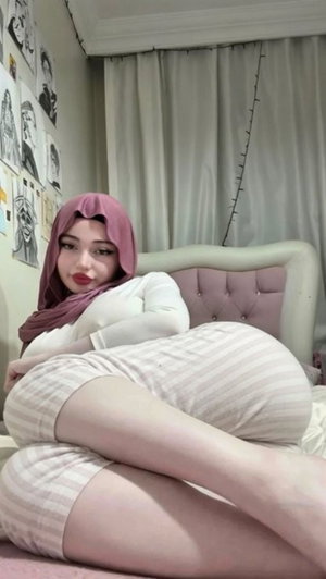 Woman in pink hijab and striped pajamas sitting on bed in bedroom.