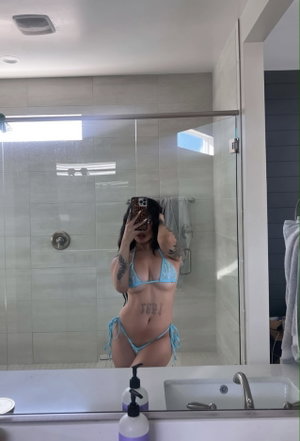 Woman in blue bikini taking mirror selfie in bathroom, tattoos visible on arms and stomach.