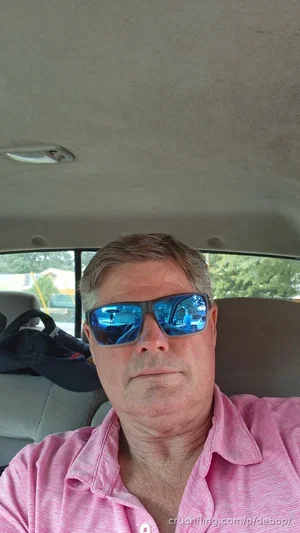 Man in pink shirt and sunglasses sitting in car, looking at camera.