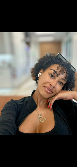 Woman with curly hair wearing black top and jacket, posing indoors with hand on chin.