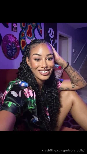 Woman with braided hair and tattoos wearing floral top, smiling in indoor setting.