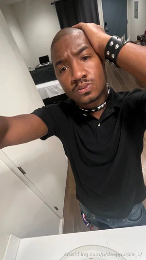 Man in black polo shirt and jeans, posing indoors with hand on head.