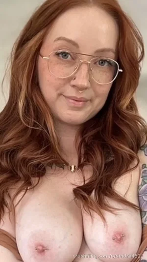 Woman with red hair and glasses, bare-chested, wearing a choker, indoors.