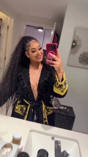 Woman with long dark hair in black and gold robe taking mirror selfie in bathroom.