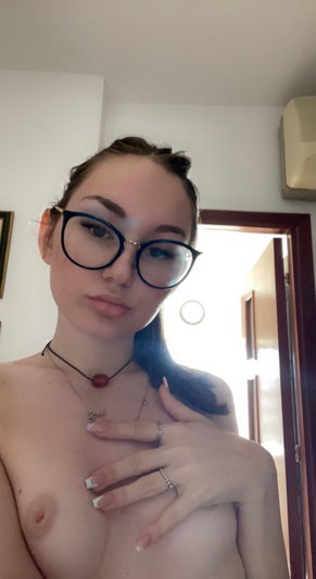 Brunette woman with glasses and necklaces posing indoors.