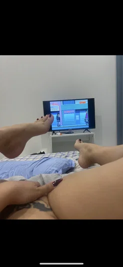 Woman lying on bed, legs up, watching TV, hand on thigh, dark nail polish.