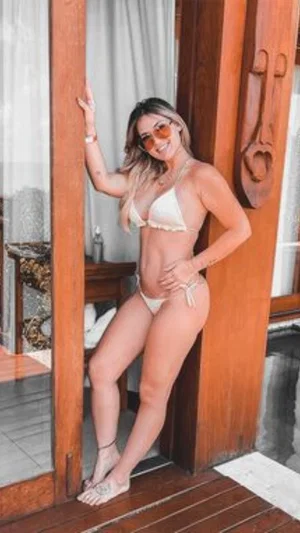Woman in white bikini posing by wooden door indoors, wearing sunglasses.