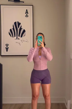 Woman in pink top and purple shorts takes mirror selfie in bedroom.