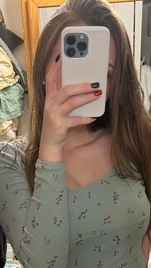 Woman with long brown hair in floral top taking mirror selfie indoors.