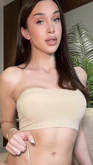 Woman with long brown hair wearing beige strapless top, sitting indoors near plant.