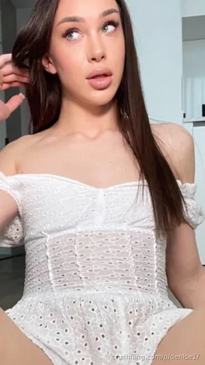 Woman with long dark hair in white off-shoulder lace dress, sitting indoors.