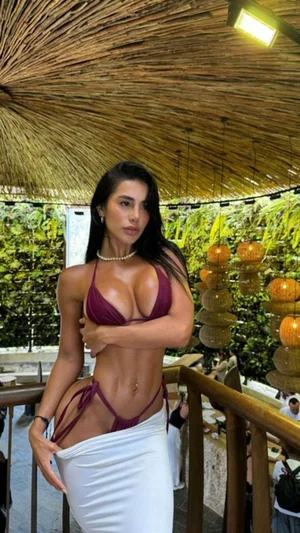 Woman in maroon bikini and white skirt posing outdoors under thatched roof.