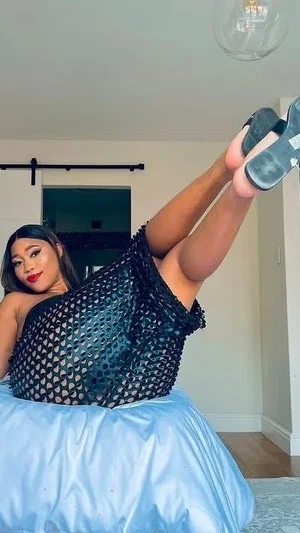 Woman in black mesh top and blue skirt posing indoors with legs raised.
