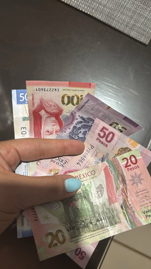 Hand with blue nails holding Mexican pesos on table.