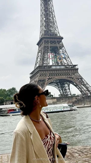Woman in beige blazer and patterned top, standing by Seine River with Eiffel Tower in background.