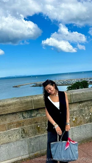 Woman in black vest and white top stands by stone wall overlooking marina under blue sky.