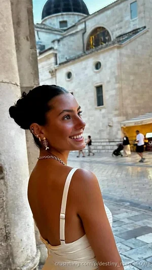 Woman in white dress, dark hair in bun, smiling, standing near stone column in outdoor plaza.