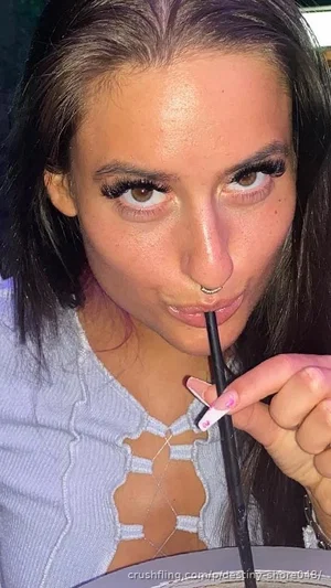 Woman with dark hair and makeup, wearing a light blue cutout top, holding a straw to her lips.