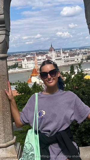 Woman with blue-tipped hair in a purple t-shirt, smiling outdoors with cityscape in background.