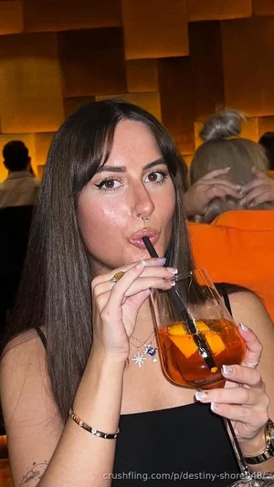 Woman with long dark hair in black top sipping drink with straw in dimly lit indoor setting.