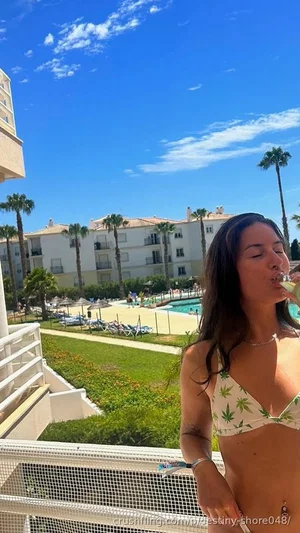Woman in bikini top on balcony, holding drink, with pool and palm trees in background.