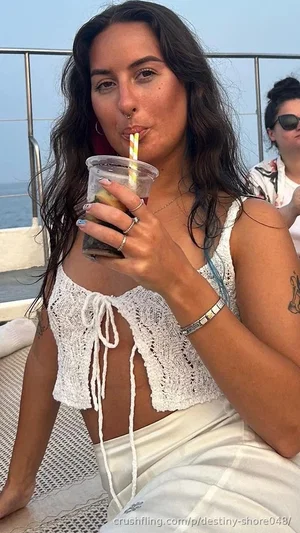 Woman with dark hair in white crocheted top and white pants, sipping drink on boat.