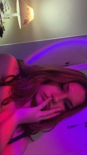 Redhead woman with hands on face in purple-lit bedroom.