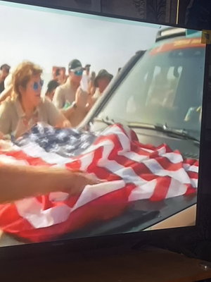 Woman with blonde hair and sunglasses holding American flag near vehicle, outdoors with crowd.
