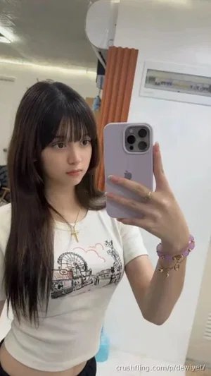 Woman with long dark hair in white graphic tee takes mirror selfie indoors.