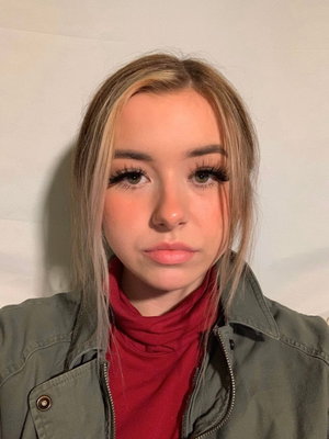 Woman with blonde hair wearing red turtleneck and green jacket, indoor selfie.