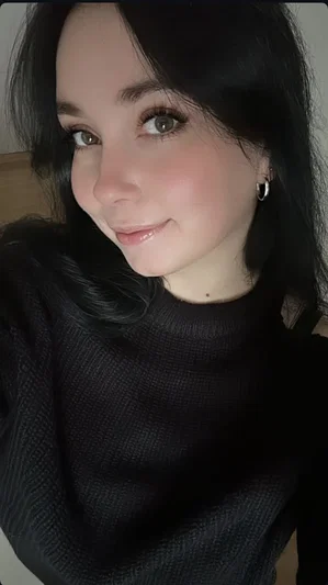 Woman with dark hair and green eyes wearing a black turtleneck, smiling softly indoors.