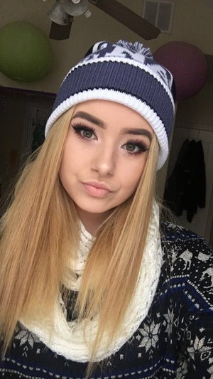 Blonde woman in winter hat and scarf, indoor selfie, cozy room setting.