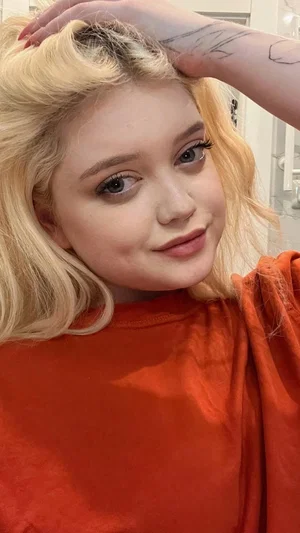 Blonde woman in orange top, hand in hair, indoor setting.